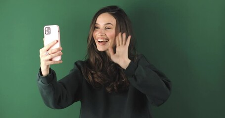 Wall Mural - Happy brunette woman having mobile video call, wave hands and say hi, hello isolated on green background. Nice to see you. Happy emotions. Beautiful young girl speaking video call online smartphone.