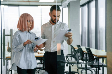 Standing and reading the document. Man and woman are working in the office together