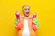 © Богдан Маліцький - crazy funny old granny in sports colorful clothes screams and shows a rock gesture on a yellow isolated background, elderly woman in youth hipster clothes