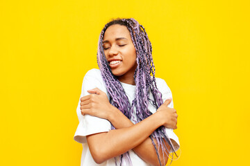 Wall Mural - young african american woman with dreadlocks hugs herself over yellow isolated background, girl with unique hairstyle and colored braids loves herself