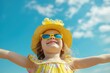 A delighted little girl wearing a yellow hat and sunglasses with arms wide open, basking in the sun.