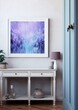 Abstract painting in purple and blue hues in a white frame on a white wall above a white console with a pink lamp and a vase on it.