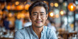 A poised Asian man, wearing glasses and a casual button-up, exudes confidence as he sits in a warmly lit restaurant, the bokeh lights complementing his stylish demeanor
