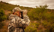 Ranger in camouflage suit and mask watches through binoculars in wooded area.