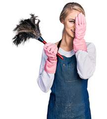 Poster - Beautiful young blonde woman wearing apron holding cleaning duster covering one eye with hand, confident smile on face and surprise emotion.