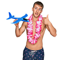 Poster - Young blond man wearing swimsuit and hawaiian lei holding airplane toy smiling happy and positive, thumb up doing excellent and approval sign