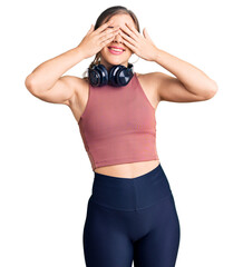 Wall Mural - Beautiful caucasian young woman wearing gym clothes and using headphones covering eyes with hands smiling cheerful and funny. blind concept.