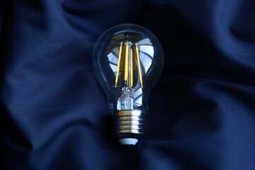 Modern quality transparent LED filament lamp with yellow filaments on dark blue silk fabric. Saving electricity. Place for the inscription. Photo. Copy space