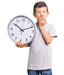 Poster - Cute blond kid holding big clock covering mouth with hand, shocked and afraid for mistake. surprised expression