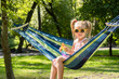 A cute five-year-old girl in a pink dress is having a great time at the park on a sunny day. She is seen blissfully relaxing in a hammock and playing with a pop it toy, surrounded by lush greenery.