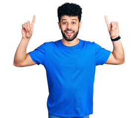 Canvas Print - Young arab man with beard wearing casual blue t shirt smiling amazed and surprised and pointing up with fingers and raised arms.