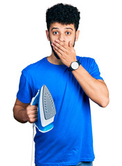 Wall Mural - Young arab man with beard holding electric steam iron covering mouth with hand, shocked and afraid for mistake. surprised expression