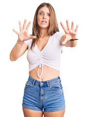 Wall Mural - Young beautiful blonde woman wearing casual white tshirt afraid and terrified with fear expression stop gesture with hands, shouting in shock. panic concept.