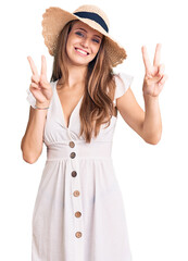 Wall Mural - Young beautiful blonde woman wearing summer dress and hat smiling looking to the camera showing fingers doing victory sign. number two.