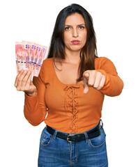 Poster - Beautiful hispanic woman holding south african 50 rand banknotes pointing with finger to the camera and to you, confident gesture looking serious