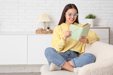 Wall Mural - Beautiful young happy Asian woman in stylish eyeglasses with book sitting on sofa at home