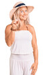 Middle age fit blonde woman wearing summer hat showing and pointing up with finger number one while smiling confident and happy.