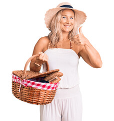 Wall Mural - Middle age fit blonde woman wearing summer hat and holding picnic wicker basket with bread smiling happy and positive, thumb up doing excellent and approval sign