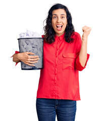 Poster - Young beautiful hispanic woman holding paper bin full of crumpled papers screaming proud, celebrating victory and success very excited with raised arms