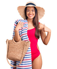 Wall Mural - Young beautiful latin girl wearing swimwear and summer hat holding beach towel and bag pointing finger to one self smiling happy and proud