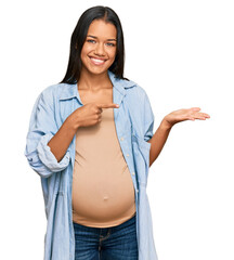 Sticker - Beautiful hispanic woman expecting a baby showing pregnant belly amazed and smiling to the camera while presenting with hand and pointing with finger.