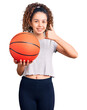 Beautiful kid girl with curly hair holding basketball ball smiling happy and positive, thumb up doing excellent and approval sign