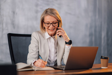 Smiling mature businesswoman talking on phone and writing in agenda.