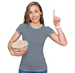 Sticker - Young blonde woman eating popcorn surprised with an idea or question pointing finger with happy face, number one