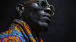 The shadows dance across the face of a smirking black man accentuating his sharp cheekbones and chiseled jawline. His vibrant printed scarf and chunky glasses add a touch of personality .