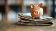 A ceramic piggy bank sits atop a stack of newspapers on a wooden surface with a blurred background