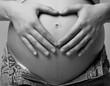 Black and white photo of  female on 6th month of pregnancy. Woman touching with hands her naked belly and shows heart sign.