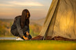 Woman sets up tent during in nature with sunset background. A method of attaching a sling when setting up a tent. Peg anchor in ground with strong fastening tent. Protecting tent from wind and rain.