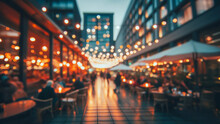 Blurred Image Shows Bokeh Of Lights Of Restaurants On Both Sides Of A Street. A Promenade In A City. Unknown People Sit At Tables By Candlelight. In The Background Is A Skyscraper.