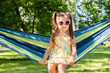 Adorable 5 years old girl with pigtails is lounging in sunglasses sitting in a hammock in a peaceful park setting on a bright, sunny day.