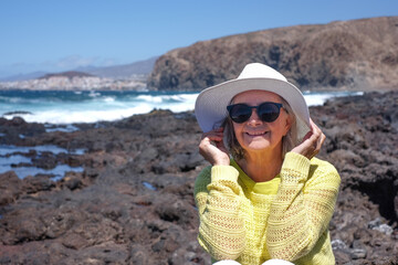 Sticker - Smiling gray haired woman on a rocky beach looking at ocean waves crash on cliffs enjoying a sunny windy day, vacation or retirement lifestyle. Horizon over water, blue sky