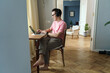  A focused man in a pink tee works on a laptop at a wooden table, headphones on, in a spacious room with classical decor.