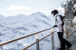 Winter sports enthusiast in Murren, Switzerland dressed in white jacket, black pants and carrying backpack overlooks snow covered mountains under cloudy skies.