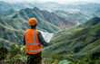 © anitjaya - An Chinese engineer wearing a yellow vest and white helmet is holding blueprints in his hand, looking at the forest mountain range on both sides of him