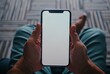 © Bambalino Studio - A person is holding a cell phone with a white screen. The phone is in their hand and they are sitting on a couch. Mockup.