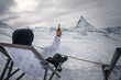 Winter scene at Zermatt ski resort person in snow gear holds up beer in deck chair overlooking Matterhorn mountain. Perfect blend of adventure and relaxation.