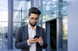 © Liubomir - Close-up photo of a young Indian man in a business suit walking on the street near an office center and using a mobile phone.