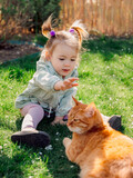 Fototapeta Do akwarium - Child girl with ginger cat on lawn in spring backyard garden