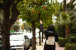 A young girl reaching up to pick a ripe orange from a tree in the bustling Athens street.