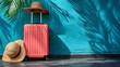 © amixstudio - Travel concept with a pink suitcase, two straw hats, and tropical palm shadow on a vibrant blue wall, suggesting a sunny vacation getaway.