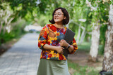 Fototapeta Mapy - A smiling Asian woman with glasses holding a laptop in a sunny park. Happy entrepreneur enjoying remote work amidst green trees outdoors