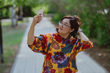 Fototapeta Zwierzęta - Joyful female capturing a selfie on a sunny day in the park. Happy woman taking a selfie with her mobile phone in a green city park