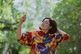 Fototapeta Zwierzęta - Cheerful young lady with glasses capturing a selfie in a lush green park. Young entrepreneur with digital devices capturing a selfie, staying connected in an urban green space