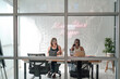 Two women working in a contemporary office, viewed through glass windows, with a 'Make Ideas Happen' neon sign. Multiethnic collaboration, focus and innovation