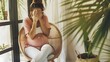 Pregnant woman in a pink shirt and white pants sitting on a tan chair with hands covering her face, feeling pain