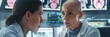 In a modern lab setting, a bald woman in a white coat speaks intently to another woman, with medical screens in the background